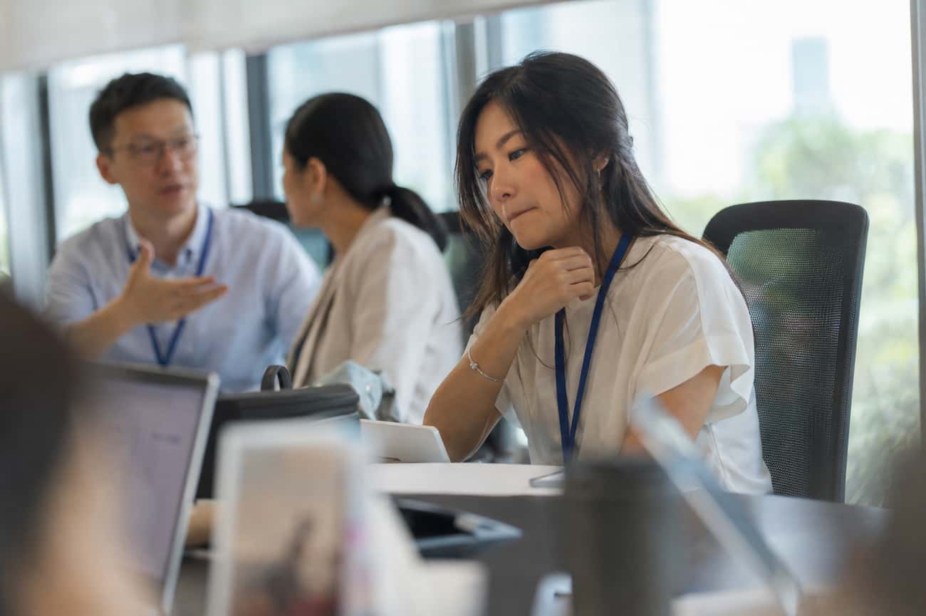A professional woman in a meeting, fist raised to her chin, looking down in quiet hesitation while a colleague speaks in the background — illustrating the experience of freezing under pressure in a workplace setting.