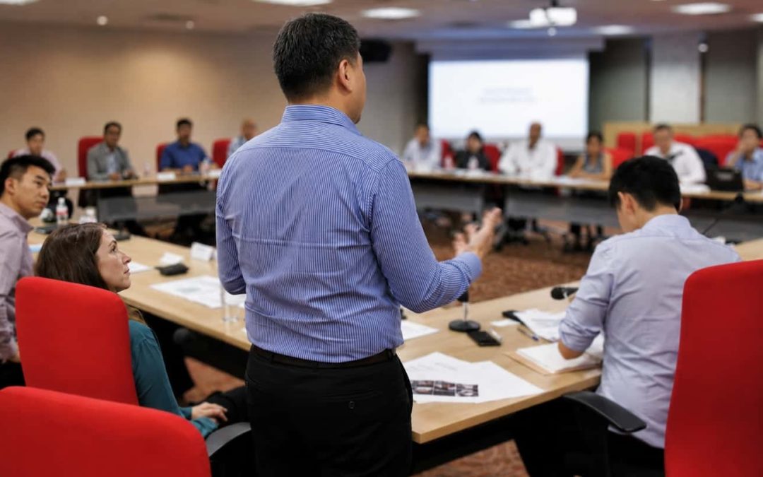 Professional man standing and presenting to a large corporate conference room, illustrating the high-stakes challenge of developing public speaking skills